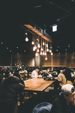 A dimly lit, cozy café filled with people sitting at long wooden tables. Warm hanging light bulbs create a relaxed atmosphere. Several individuals are engaged with their laptops and phones, suggesting a busy yet peaceful environment.