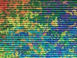 A corrugated metal surface is painted with a vibrant camouflage pattern. The colors blend into each other in an abstract manner, featuring shades of green, blue, yellow, orange, and purple. The paint creates an intricate and dynamic visual texture.