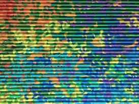 A corrugated metal surface is painted with a vibrant camouflage pattern. The colors blend into each other in an abstract manner, featuring shades of green, blue, yellow, orange, and purple. The paint creates an intricate and dynamic visual texture.