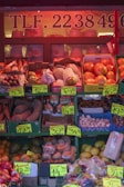 A market stall displaying a variety of fruits and vegetables with bold price signs. The produce includes oranges, bananas, sweet potatoes, mushrooms, and more, packed in crates and boxes. The stall is lit with a warm reddish tone, creating a cozy and inviting atmosphere.