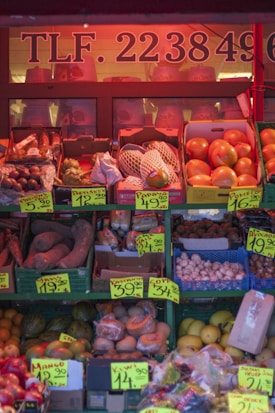 A market stall displaying a variety of fruits and vegetables with bold price signs. The produce includes oranges, bananas, sweet potatoes, mushrooms, and more, packed in crates and boxes. The stall is lit with a warm reddish tone, creating a cozy and inviting atmosphere.