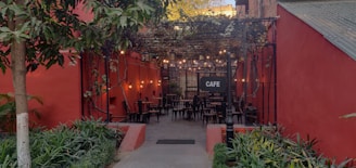 An outdoor cafe with red walls and a cozy atmosphere. The seating area features multiple black chairs and tables under a canopy of vines, with string lights adding a warm glow. Surrounding the entrance are green plants and a pathway leading inside.