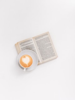 A steaming cup of coffee with a detailed face printed on the foam, set beside an open book