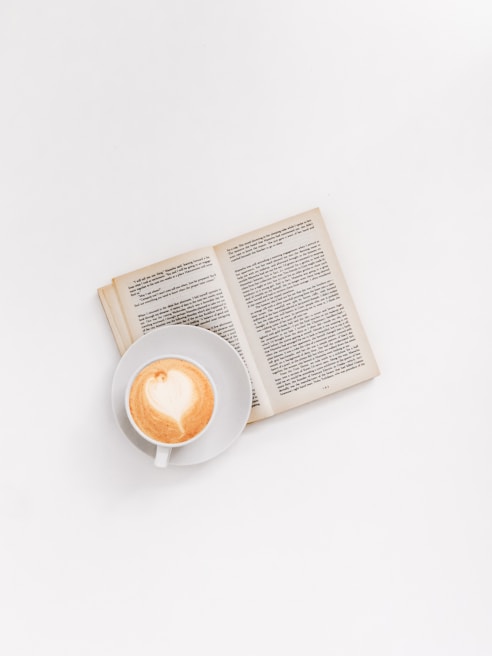 A steaming cup of coffee with a detailed face printed on the foam, set beside an open book