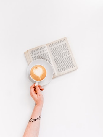 A hand with a tattoo of a branch holds a coffee cup with latte art. An open book lies on a white background.