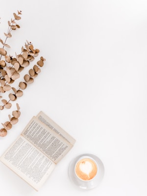 An open book with visible text placed on a white surface next to a cup of coffee with latte art in the form of a heart. Several sprigs of dried eucalyptus leaves are artistically arranged alongside.