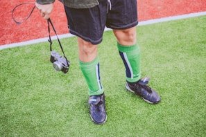 A person stands on a grassy sports field, wearing green knee-high socks and black athletic shoes. The individual is holding a camera by its strap in one hand. The field features a red and green artificial turf.