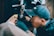 A tattoo artist is intensely focused while tattooing a person's arm. The artist is wearing a black cap and gloves, holding a tattoo machine. The background includes clothing hung up, possibly in a studio.