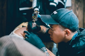 A tattoo artist is intensely focused while tattooing a person's arm. The artist is wearing a black cap and gloves, holding a tattoo machine. The background includes clothing hung up, possibly in a studio.