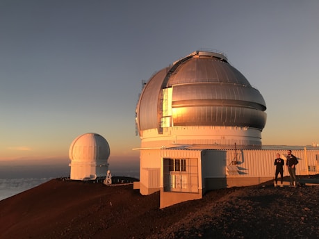 Two large observatory domes are situated on a mountain top with a clear sky at sunset. The larger dome is in the foreground while the smaller one is in the background, both reflecting the golden hues of the setting sun. Two people are standing on the right side, near the larger observatory structure.