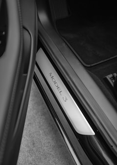 A close-up of a car interior, focusing on the door sill where 'Model 3' is displayed. The image is in black and white and shows the sleek design of the car's entrance area with clean lines and a metallic finish.