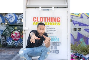 A person crouches in front of a clothing and shoe donation bin with colorful graffiti-covered walls in the background. The bin has text indicating it's for the National Children's Cancer Society. The person is wearing a black shirt and ripped jeans, pointing towards the camera. A pile of clothing items is visible on the ground nearby.