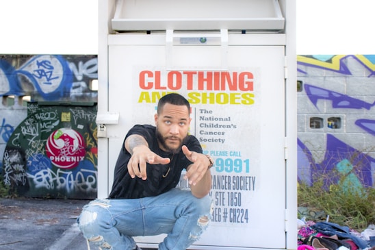A person crouches in front of a clothing and shoe donation bin with colorful graffiti-covered walls in the background. The bin has text indicating it's for the National Children's Cancer Society. The person is wearing a black shirt and ripped jeans, pointing towards the camera. A pile of clothing items is visible on the ground nearby.