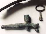 A collection of antique metal objects including a long curved piece, an old key with a round handle, and a vintage nozzle or spout. The objects have a rustic, aged appearance with a tarnished patina, set against a plain background.
