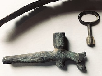 A collection of antique metal objects including a long curved piece, an old key with a round handle, and a vintage nozzle or spout. The objects have a rustic, aged appearance with a tarnished patina, set against a plain background.