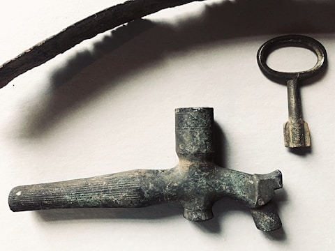 A collection of antique metal objects including a long curved piece, an old key with a round handle, and a vintage nozzle or spout. The objects have a rustic, aged appearance with a tarnished patina, set against a plain background.