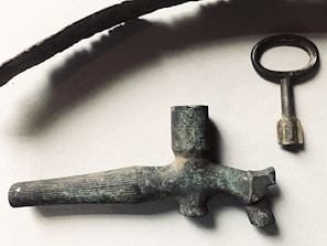 A collection of antique metal objects including a long curved piece, an old key with a round handle, and a vintage nozzle or spout. The objects have a rustic, aged appearance with a tarnished patina, set against a plain background.