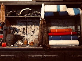 A rustic setting featuring various objects on wooden shelves against a stone wall. Blankets in white, blue, and red are neatly stacked on one side, while the other shelf holds a pirate-style hat and several black mugs. Below, an assortment of items including axes, red bottles, a fur pelt, ropes, and a wooden barrel are displayed.