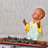 A small ceramic figure of a monk with a peaceful expression, wearing a light green robe with yellow trim, is placed on a textured background. In front of the figure is a red tray filled with dried tea leaves.