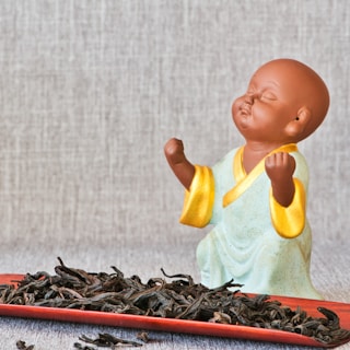 A small ceramic figure of a monk with a peaceful expression, wearing a light green robe with yellow trim, is placed on a textured background. In front of the figure is a red tray filled with dried tea leaves.