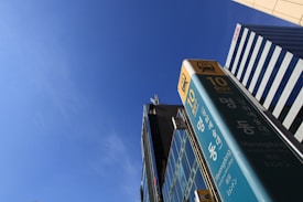 A tall signpost with Korean characters stands prominently against a clear blue sky. The sign includes the number '10' and the name 'Myeongdong' in multiple languages. In the background, modern buildings with striped patterns extend upward.