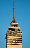 A tall skyscraper with a digital clock displaying the time 17:36:09 and the words 'LATINO AMERICANA' prominently displayed. The building structure has multiple antennas and a clear design with windows and horizontal bands. The clear blue sky provides a backdrop, enhancing the architectural detail and symmetry.
