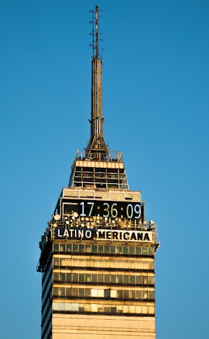 A tall skyscraper with a digital clock displaying the time 17:36:09 and the words 'LATINO AMERICANA' prominently displayed. The building structure has multiple antennas and a clear design with windows and horizontal bands. The clear blue sky provides a backdrop, enhancing the architectural detail and symmetry.