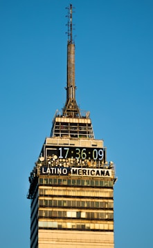 A tall skyscraper with a digital clock displaying the time 17:36:09 and the words 'LATINO AMERICANA' prominently displayed. The building structure has multiple antennas and a clear design with windows and horizontal bands. The clear blue sky provides a backdrop, enhancing the architectural detail and symmetry.