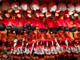 A collection of plush toys arranged in rows, featuring characters dressed in red and white outfits with green scarves and black boots, reminiscent of Santa Claus. The toys are hanging from above, creating a colorful and festive display.