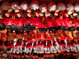A collection of plush toys arranged in rows, featuring characters dressed in red and white outfits with green scarves and black boots, reminiscent of Santa Claus. The toys are hanging from above, creating a colorful and festive display.