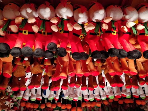 A collection of plush toys arranged in rows, featuring characters dressed in red and white outfits with green scarves and black boots, reminiscent of Santa Claus. The toys are hanging from above, creating a colorful and festive display.