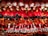 A collection of plush toys arranged in rows, featuring characters dressed in red and white outfits with green scarves and black boots, reminiscent of Santa Claus. The toys are hanging from above, creating a colorful and festive display.