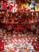 A vibrant display of various plush toys, mostly arranged in rows and piled up at the bottom. The toys include teddy bears, snowmen, and other festive characters, with prominent red, white, brown, and black colors. The arrangement has a festive and playful appearance, suggesting a carnival or amusement park setting.