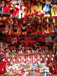 A vibrant display of various plush toys, mostly arranged in rows and piled up at the bottom. The toys include teddy bears, snowmen, and other festive characters, with prominent red, white, brown, and black colors. The arrangement has a festive and playful appearance, suggesting a carnival or amusement park setting.