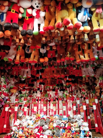 A vibrant display of various plush toys, mostly arranged in rows and piled up at the bottom. The toys include teddy bears, snowmen, and other festive characters, with prominent red, white, brown, and black colors. The arrangement has a festive and playful appearance, suggesting a carnival or amusement park setting.
