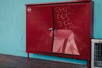 A red metal cabinet with a dent on its surface is positioned against a teal wall. The cabinet features graffiti with white scribbles and has a handle on one side. The floor is made of brownish tiles, and part of a metal vent or air conditioning unit is visible on the right side.