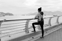 A person stands on a waterfront promenade, gazing out at a calm body of water. The scene is in black and white, with distant mountains and an urban skyline visible. The individual is wearing casual clothing, including a denim jacket and leggings, with their hair tied up in a bun.