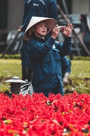 A person wearing a traditional conical hat and blue clothing is standing amidst a field of vibrant red flowers, capturing a photo with a camera. In the background, there are other individuals and green foliage.