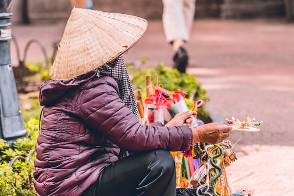 A person wearing a traditional conical hat and a dark puffy jacket is sitting on the ground, interacting with various colorful handcrafted items likely for sale. The scene includes vibrant toys and objects around them, suggesting a street market or outdoor setting. In the background, a pedestrian with white pants walks past.