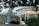 An elderly person with short gray hair sits cross-legged on a mat near a pond, deeply engrossed in reading an open book. A beige handbag is placed beside them. The background features a park setting with blurred figures sitting on benches and trees lining the pond.