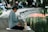 An elderly person with short gray hair sits cross-legged on a mat near a pond, deeply engrossed in reading an open book. A beige handbag is placed beside them. The background features a park setting with blurred figures sitting on benches and trees lining the pond.