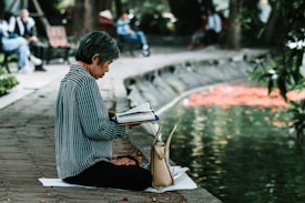 An elderly person with short gray hair sits cross-legged on a mat near a pond, deeply engrossed in reading an open book. A beige handbag is placed beside them. The background features a park setting with blurred figures sitting on benches and trees lining the pond.