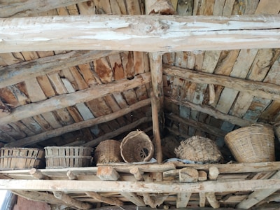 The image features a rustic wooden attic or loft area with multiple woven baskets placed on wooden beams. The ceiling is constructed from natural, unpolished wooden planks and beams, giving a traditional and aged appearance.