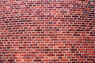 A curved brick wall features a pattern of horizontal rows of reddish-brown bricks separated by gray mortar. The bricks have varying tones of red and brown, indicating weathering and age.