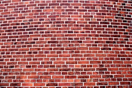 A curved brick wall features a pattern of horizontal rows of reddish-brown bricks separated by gray mortar. The bricks have varying tones of red and brown, indicating weathering and age.