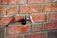 A silver outdoor faucet is mounted on a red brick wall. The bricks are weathered, showing various shades of red and brown. The faucet casts a shadow, indicating bright lighting conditions.