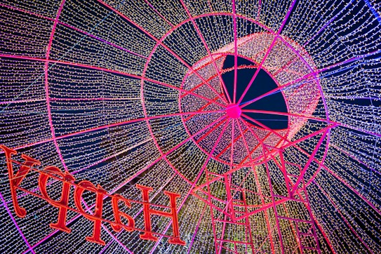 An intricate design of illuminated red and pink framework creating a mesmerizing pattern of lights, forming a canopy of twinkling fairy lights against a dark sky. The word 'HAPPY' is prominently displayed in red letters, appearing as part of the festive decorations.