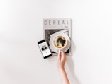 A minimalist composition featuring a hand holding a coffee cup placed on top of a magazine titled 'Cereal'. Beside it, a smartphone displays a music player with a song paused.