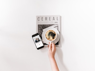 A minimalist composition featuring a hand holding a coffee cup placed on top of a magazine titled 'Cereal'. Beside it, a smartphone displays a music player with a song paused.