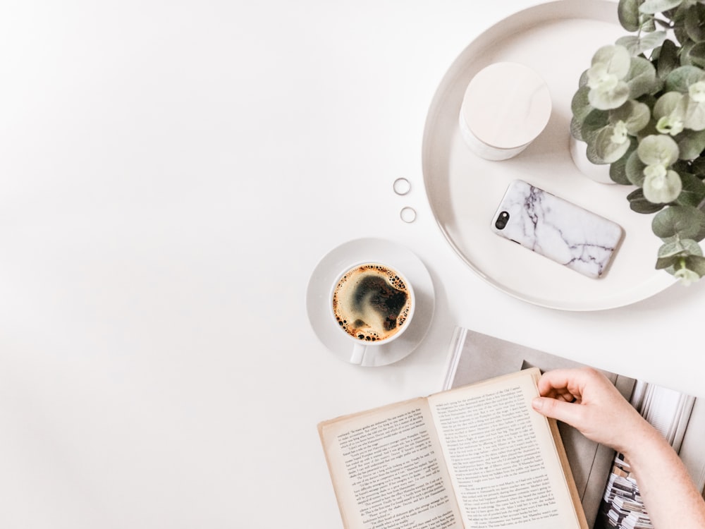 Flat-Lay-Fotografie einer Person, die ein aufgeschlagenes Buch auf der Tischplatte neben einer Tasse schwarzen Kaffees hält